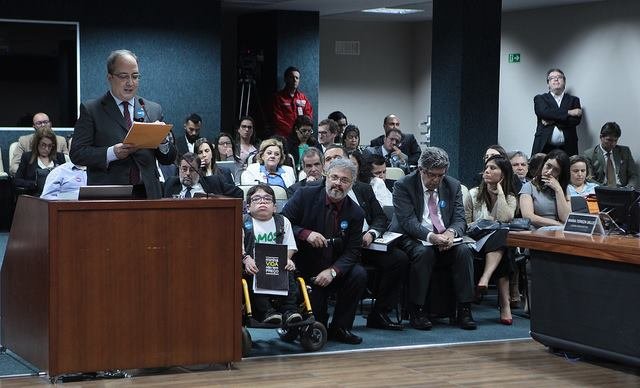 Audiencia pública con oradores y participantes - Solidaritas Hombre de pie leyendo un documento en una audiencia pública con los participantes sentados al fondo.