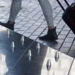 Una persona con pantalones a cuadros y botas negras tirando de una maleta flanqueada por una superficie con alfileres metálicos.