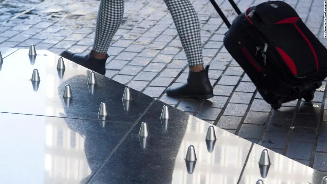 Persona caminando con maleta flanqueada por superficie con alfileres metálicos - Solidaritas Una persona con pantalones a cuadros y botas negras tirando de una maleta flanqueada por una superficie con alfileres metálicos.