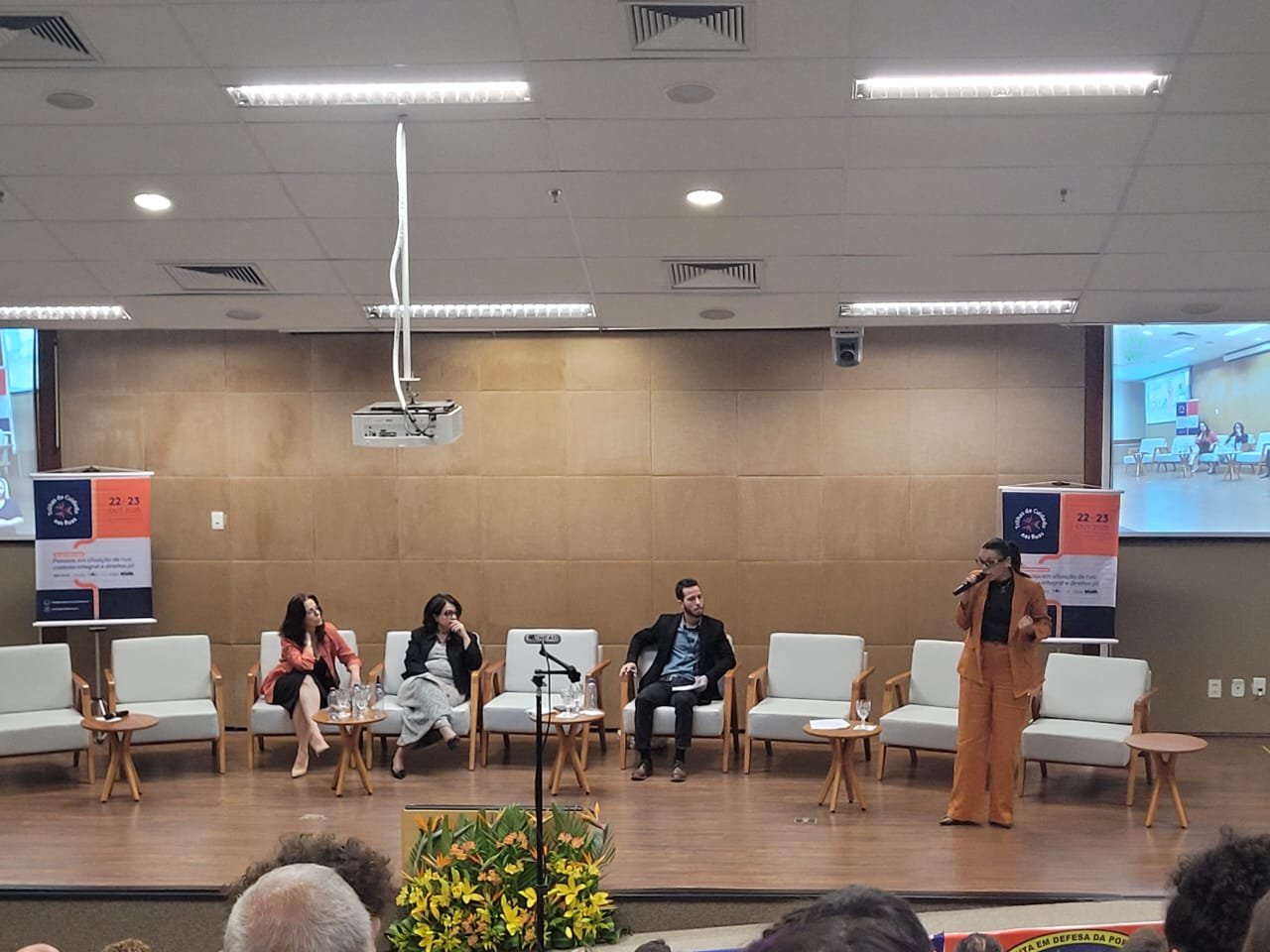 Painel de discussão em seminário com quatro pessoas em um palco e telão ao fundo.