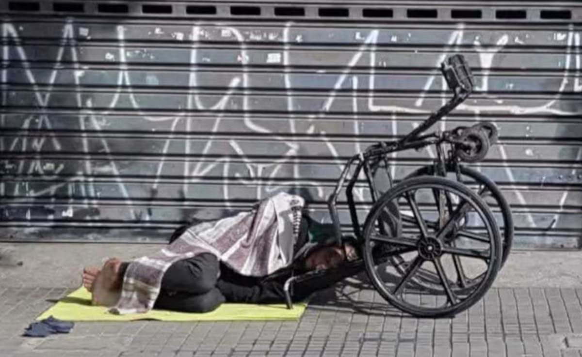 Indivíduo deitado em uma esteira com uma cadeira de rodas ao lado, coberto por um lençol.