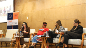 Quatro mulheres sentadas em uma mesa durante um seminário, uma delas falando ao microfone.