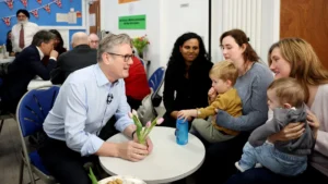 Keir Starmer sentado à mesa com uma família, segurando um vaso de flores enquanto conversa com crianças.