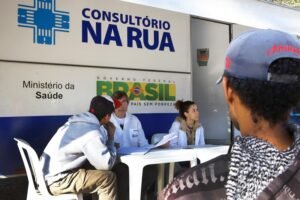 Profissionais de saúde atendendo pessoas em um consultório móvel na rua, com a marca do Ministério da Saúde do Brasil.