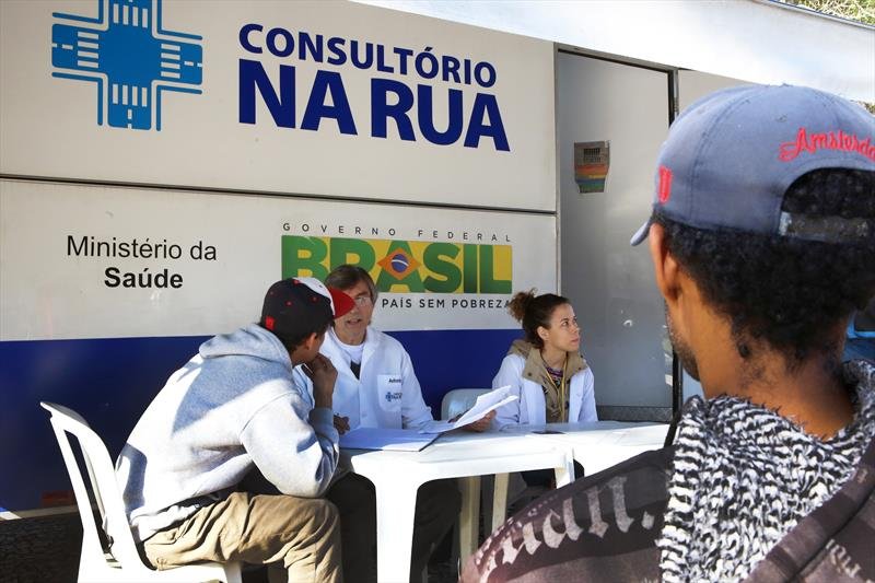 Profissionais de saúde atendendo pessoas em um consultório móvel na rua, com a marca do Ministério da Saúde do Brasil.