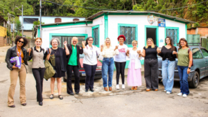 Um grupo de mulheres sorrindo e unidas em um evento comunitário, posando para a foto em frente a uma casa.